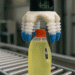 SOMARICS Robotics soft gripper with blue-tipped flexible fingers lifting a yellow detergent bottle from a conveyor in an industrial setting.